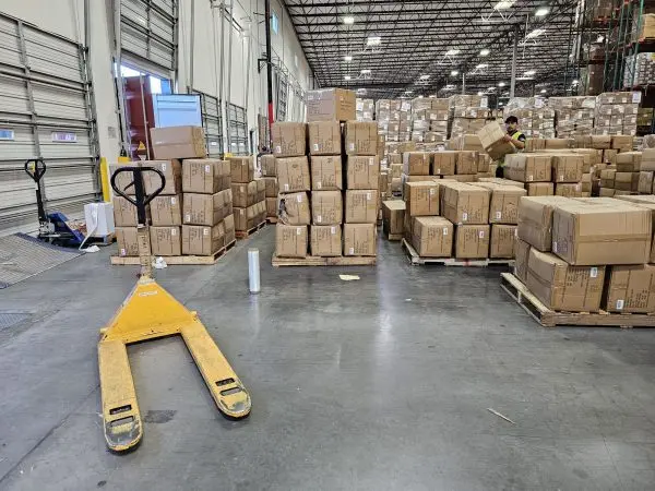 Palletized cartons staged on a warehouse floor next to a pallet jack, with a worker stacking boxes